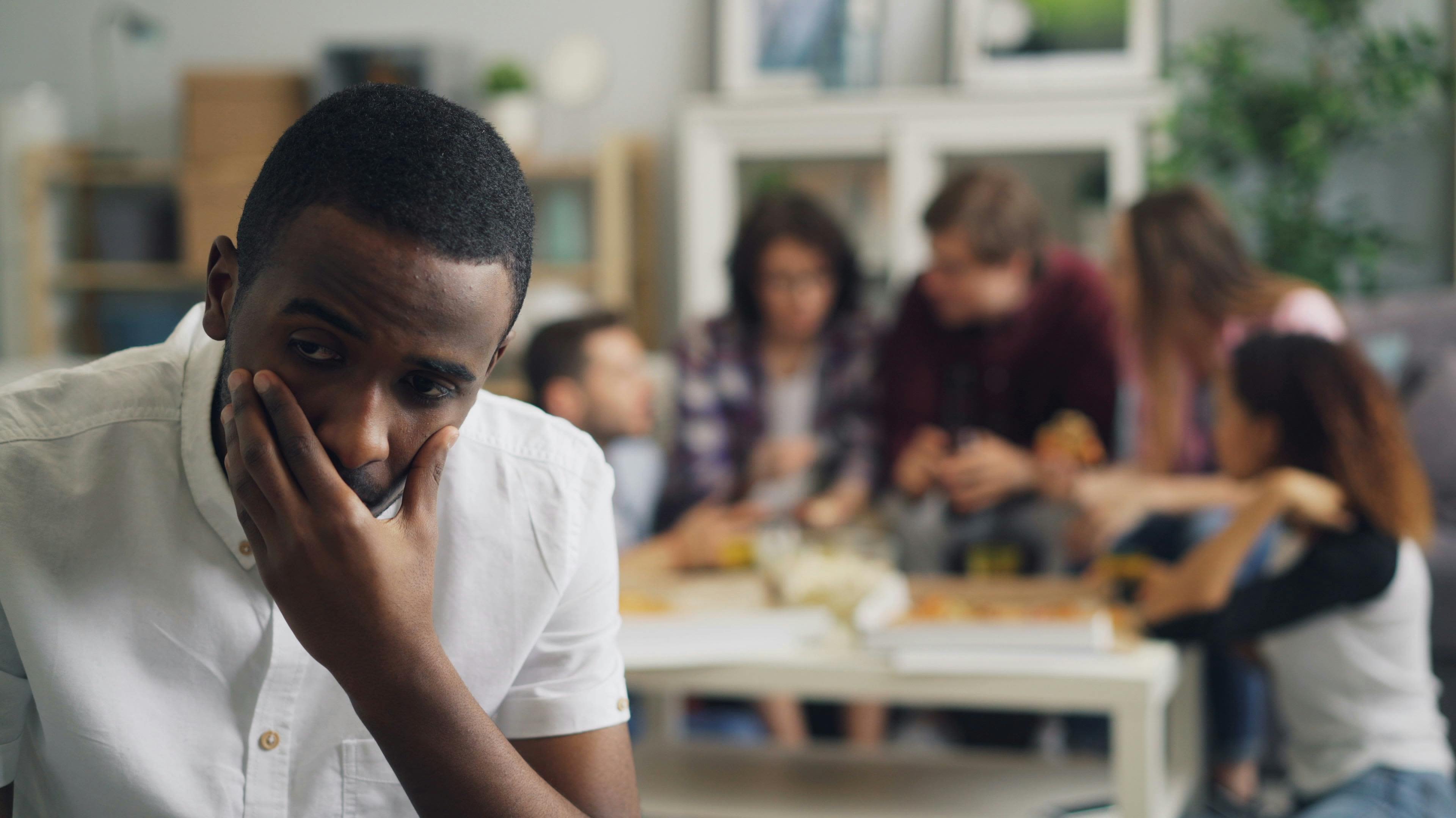 A concerned man sits apart from a lively group in a cozy home setting.- Photo by Vitaly Gariev on Pexels