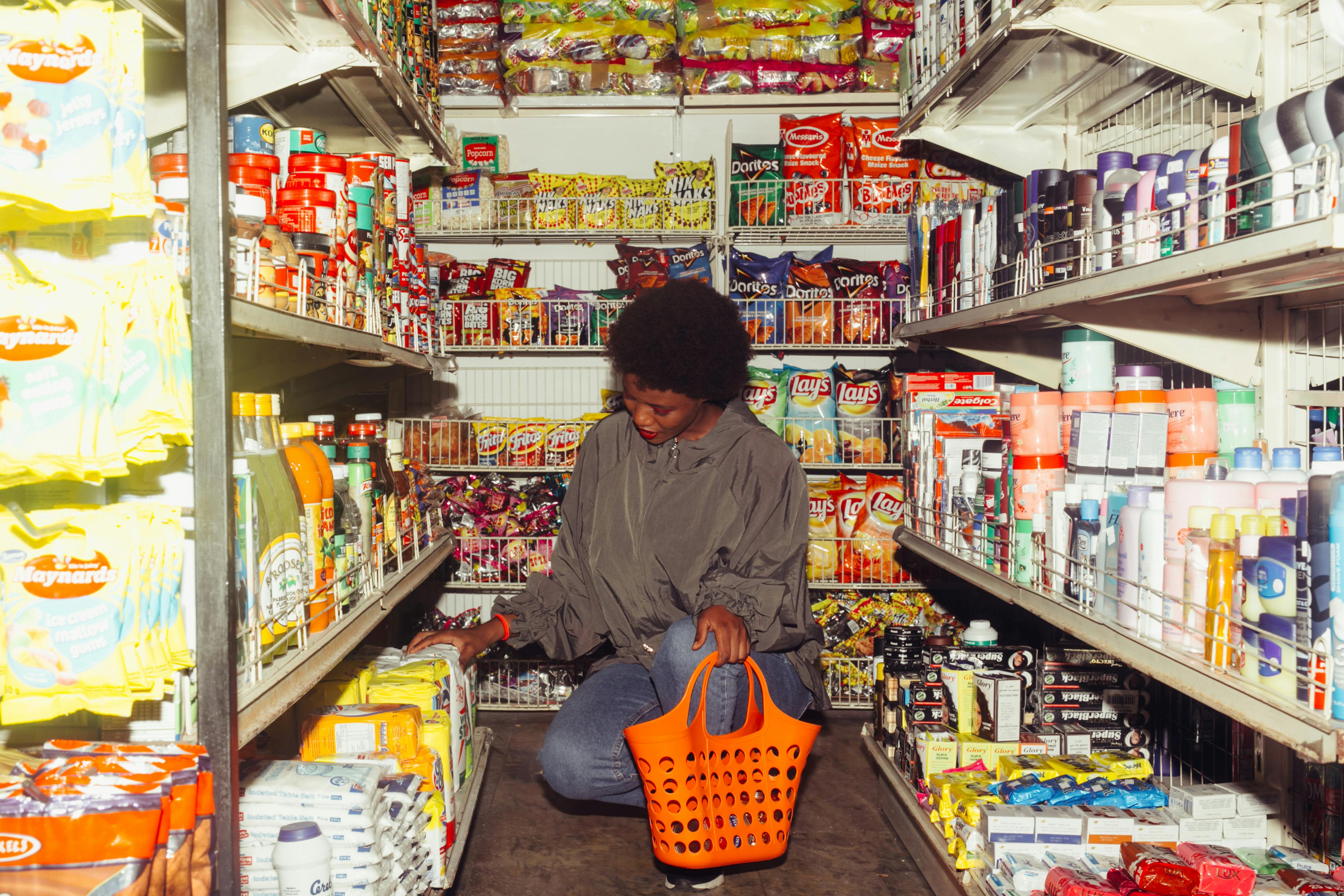 Full length of adult ethnic female buyer selecting goods while squatting with shopping bag among grocery shelves